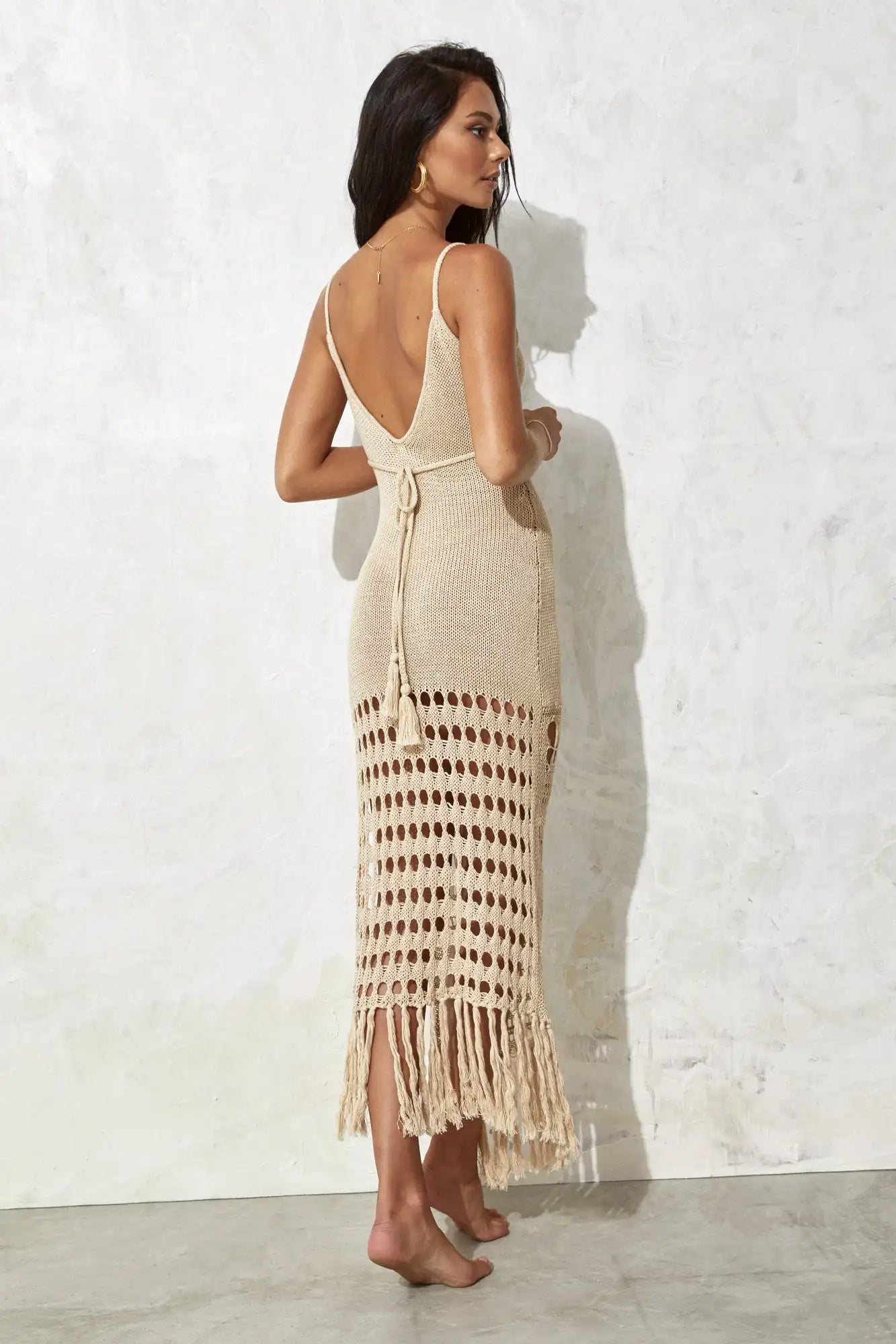 Woman wearing a beige open-back dress with fringe details against a white wall.