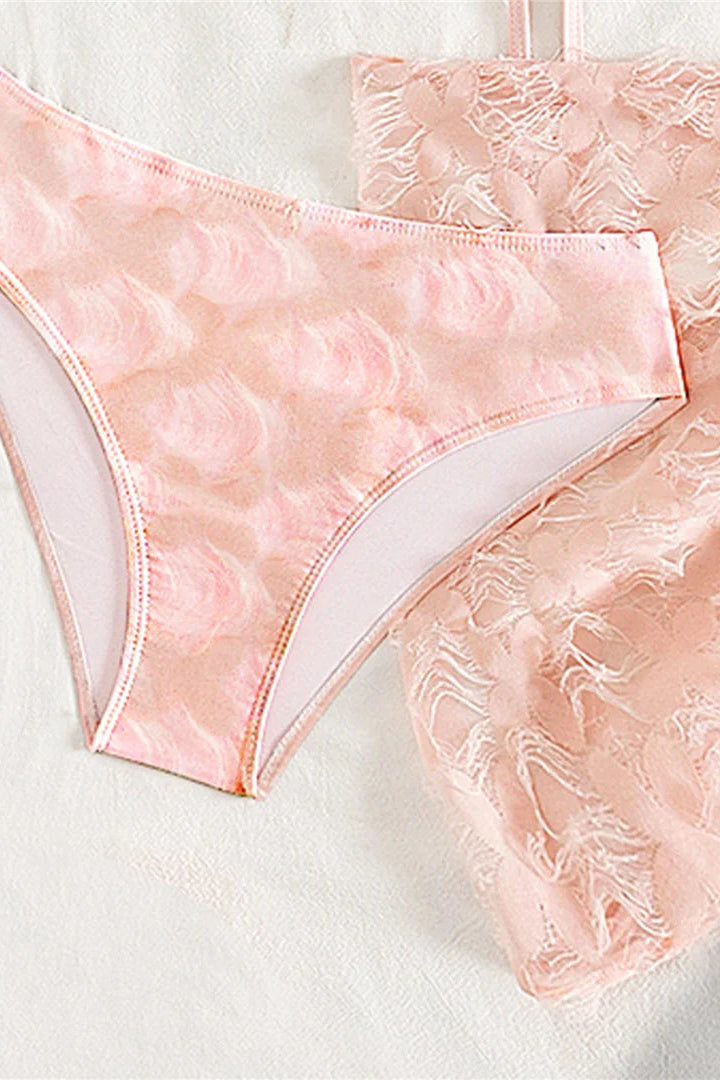 A close-up of a light pink bikini bottom with a textured, raised floral or scallop pattern, with a matching sheer sarong partially visible behind it.