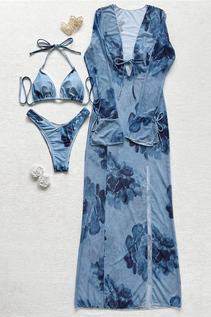 A coordinated swimwear set laid flat on a light textured background, including a long blue floral cover-up, a matching bikini top, bikini bottom