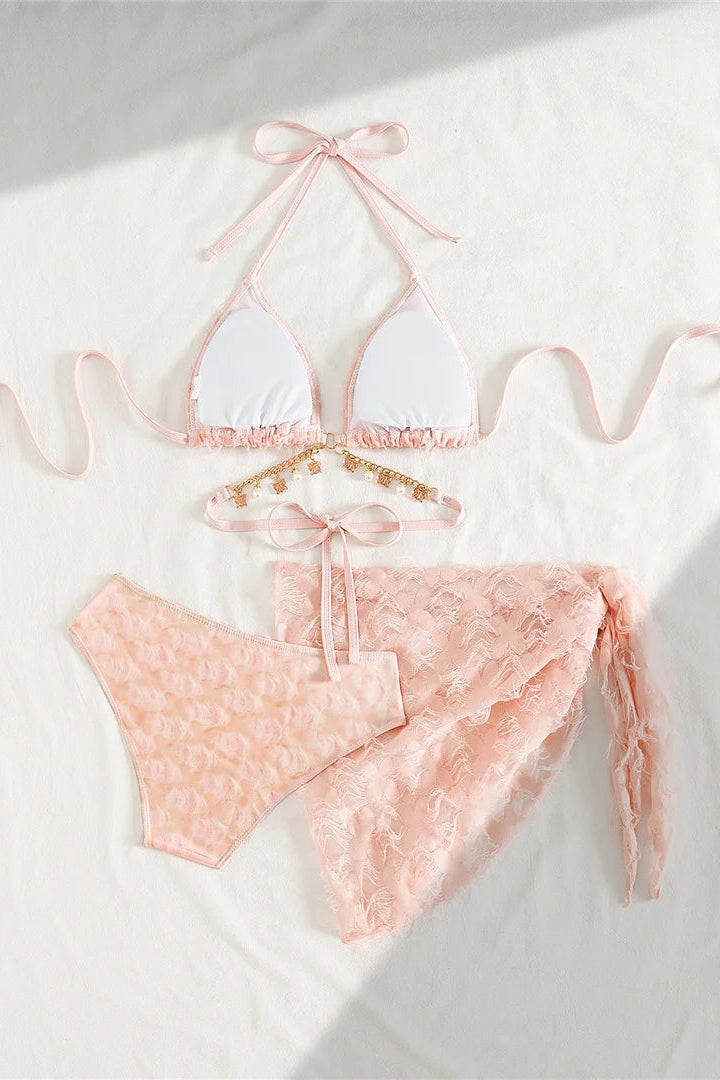 A pastel pink two-piece bikini set includes a triangle halter top with white cups matching pink bikini bottoms, a sheer pink lace wrap, and a small gold-tone beaded belt accessory.