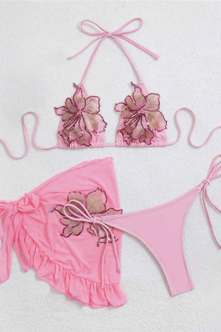 A pink two-piece bikini set with matching swim bottom. The bikini top has large floral appliques in shades of pink and beige, and the matching bottoms feature a ruffled pink wrap-style cover-up tied at the side.