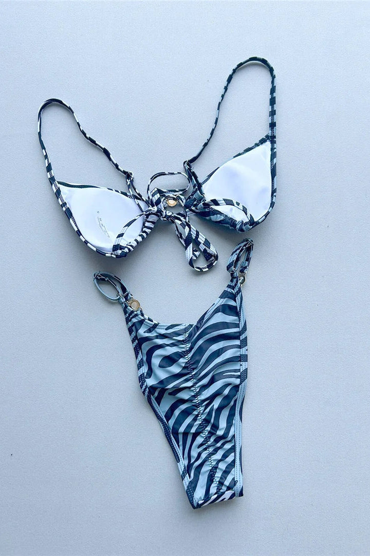 A two-piece bikini set laid flat  The bikini top has white cups with a dark blue and black abstract patterned trim and tie, and the matching high-cut bottoms feature a blue and black zebra-like pattern.