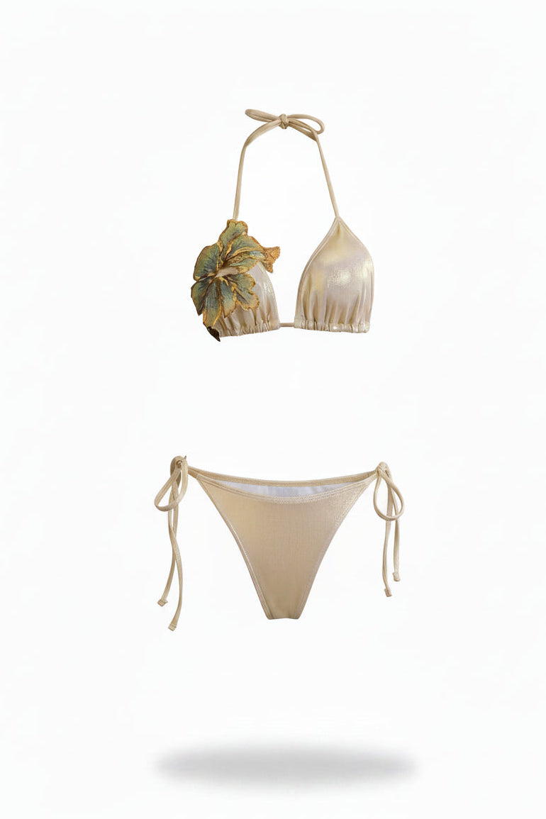 A two-piece string bikini set in a light gold or cream color, featuring a halter top with a large 3D floral applique on one cup and matching tie-side bottoms.