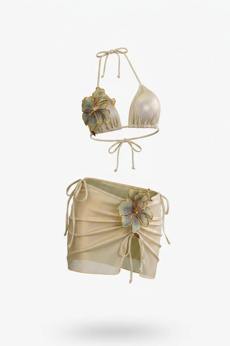 A two-piece swimwear set featuring a halter bikini top and matching wrap-style bikini bottoms cover up in a champagne beige color, both adorned with large 3D floral embellishments in gold, blue, and green, 