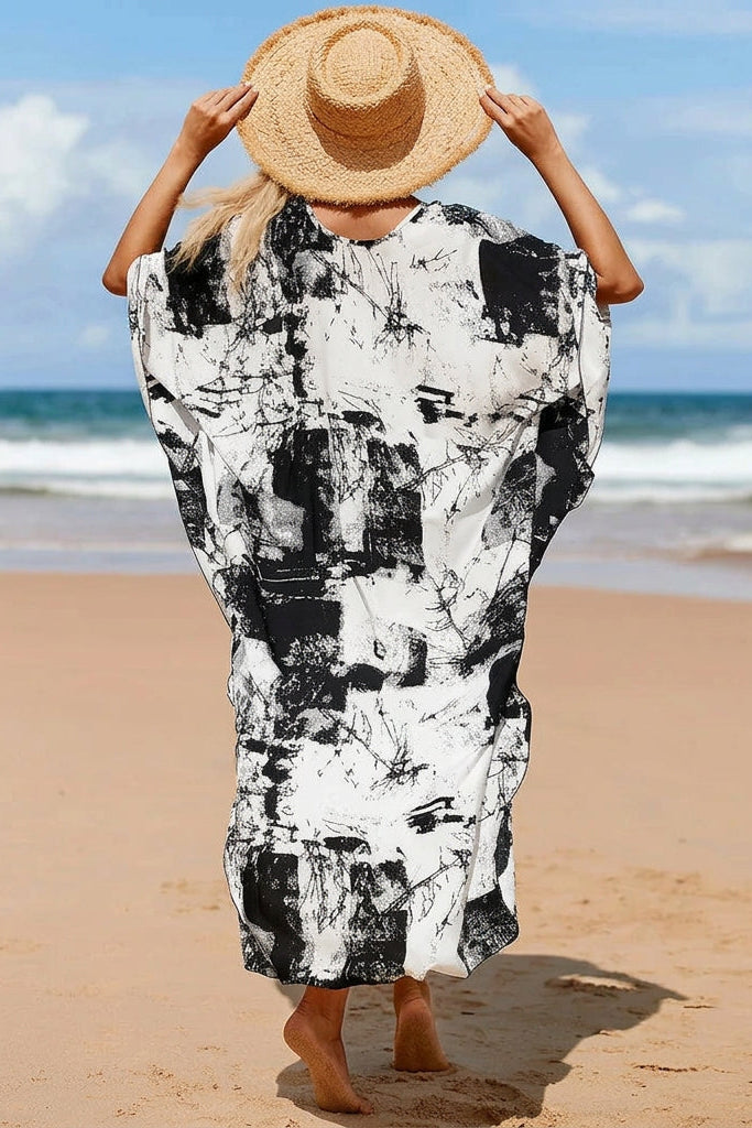A woman on a sandy beach seen from behind, wearing a loose black and white abstract print kaftan dress