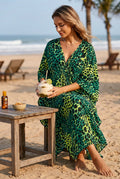 A woman on a sandy beach wearing a flowing leopard-print kaftan dress in green, black, and yellow-green tones.