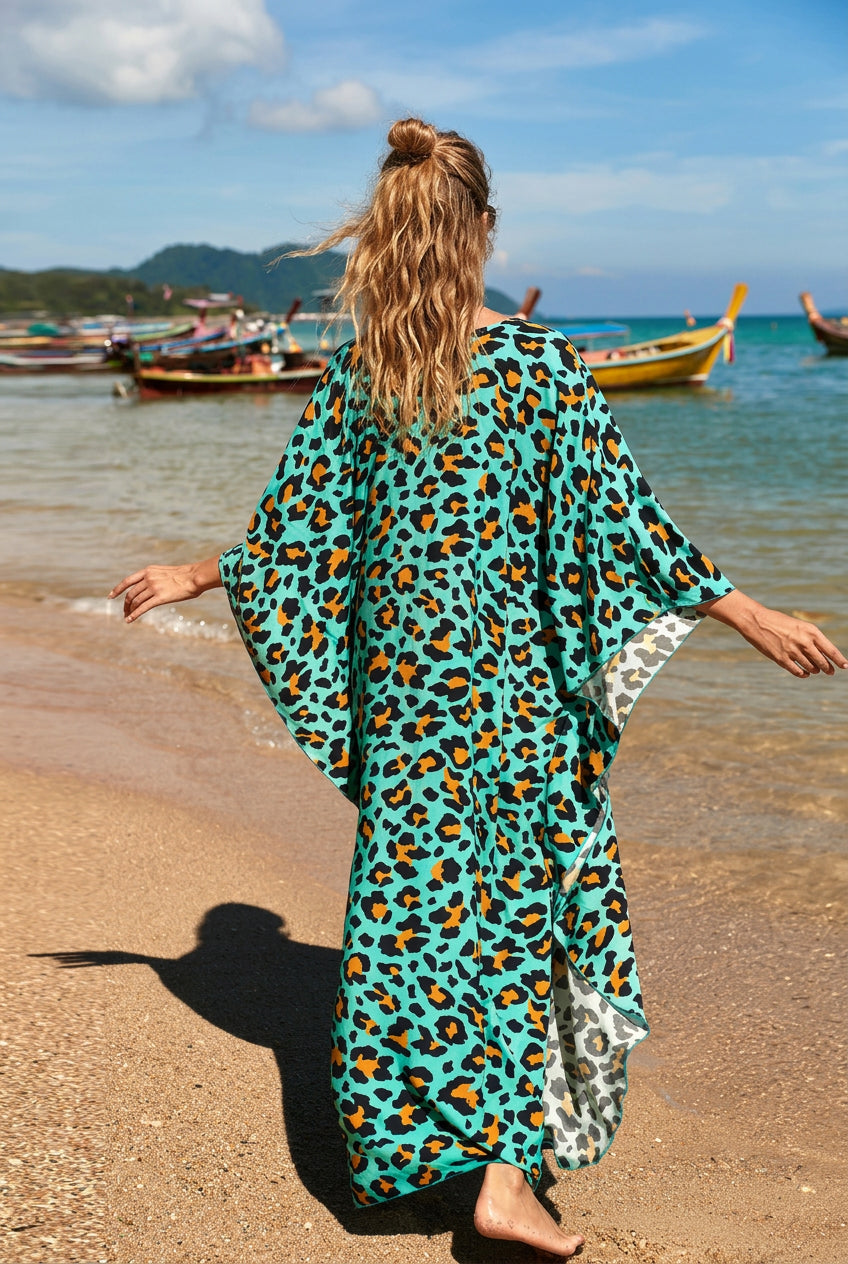A woman stands on a sandy beach facing away from the camera, wearing a flowing turquoise leopard-print kaftan dress with wide sleeves and a patterned inner lining.