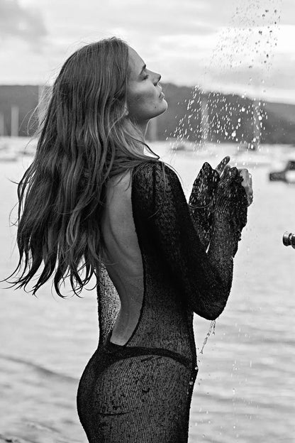 Black and white photo of a woman in a cover up dress by a window with water outside