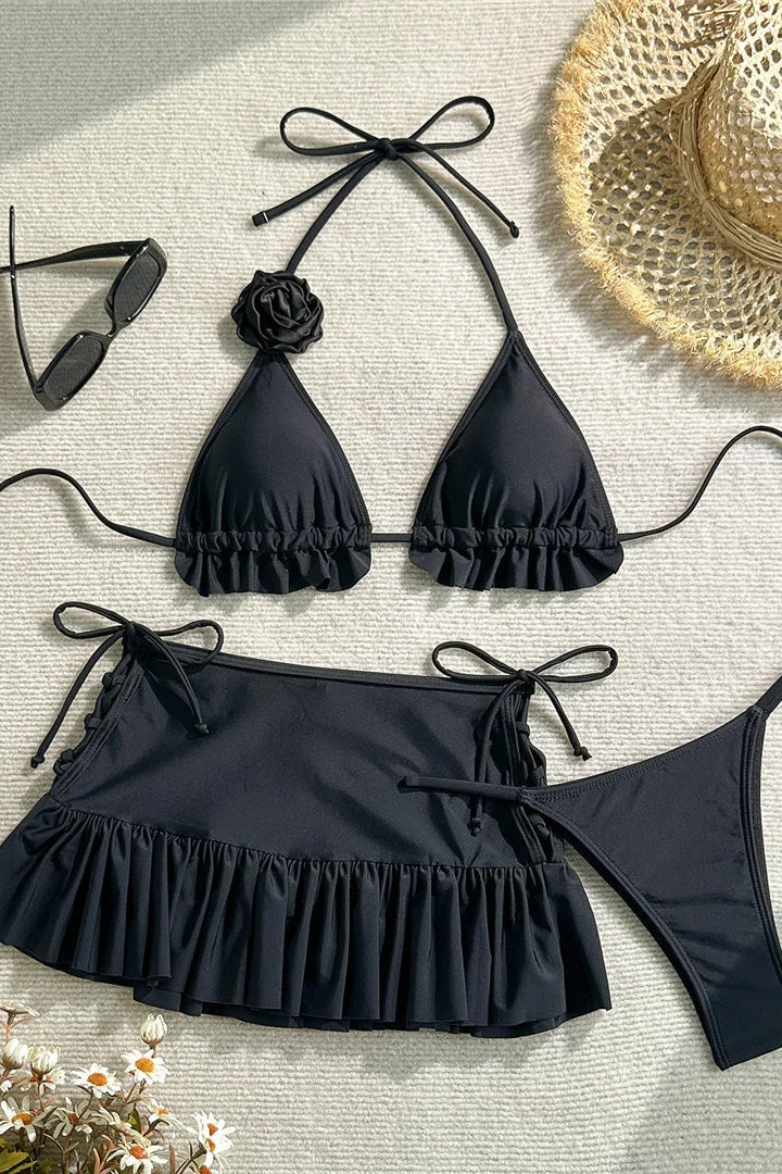 Black bikini set with ruffled details, sunglasses, and a straw hat on a light surface.