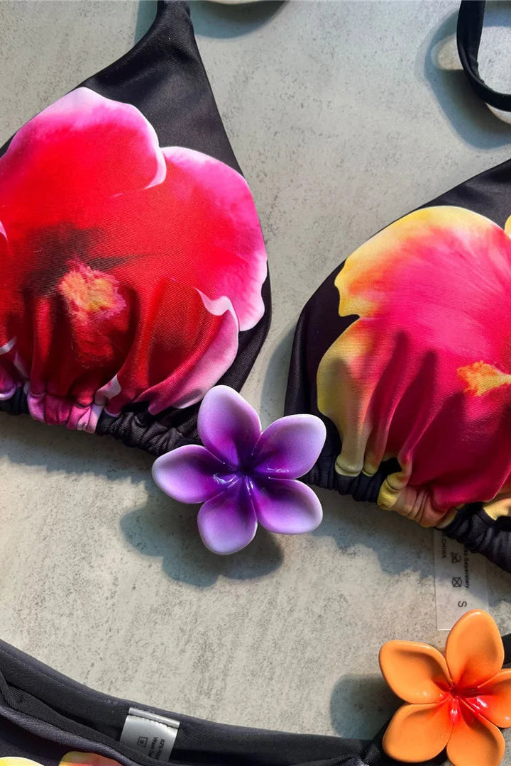 Black bikini top with large red, pink, and yellow tropical flower prints, shown on a light gray surface with small purple and orange 3D flower accent.