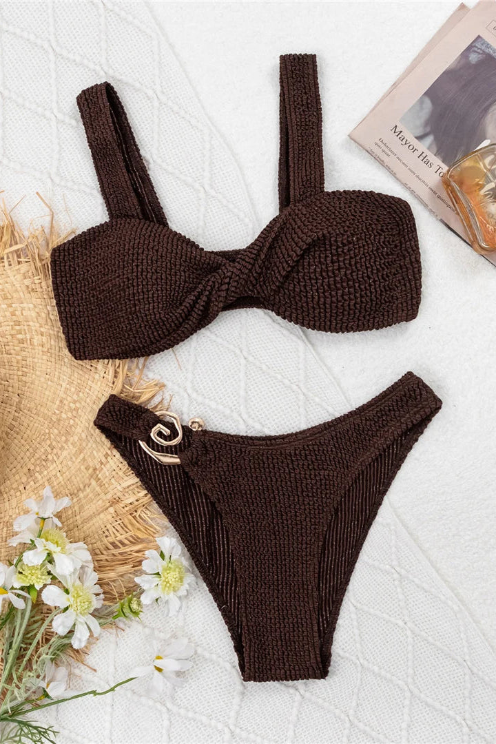 Brown knitted bikini set on a white surface with a straw hat and flowers.