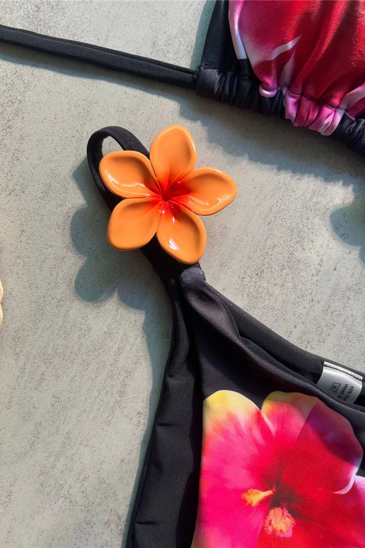 Close-up of a bikini top strap with a glossy orange plumeria flower charm, paired with a black swimsuit featuring a large pink and yellow tropical flower print, laid on a gray concrete surface.