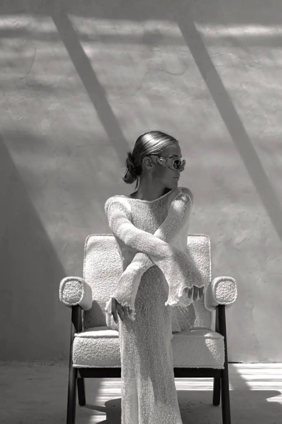 Female in a textured sheer cover up dress sitting on a chair with sunlight casting shadows.