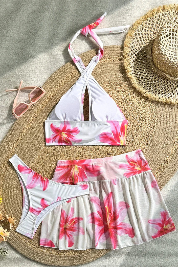 Floral bikini set with matching cover up mini skirt, straw hat, sunglasses, and flowers on a textured surface.