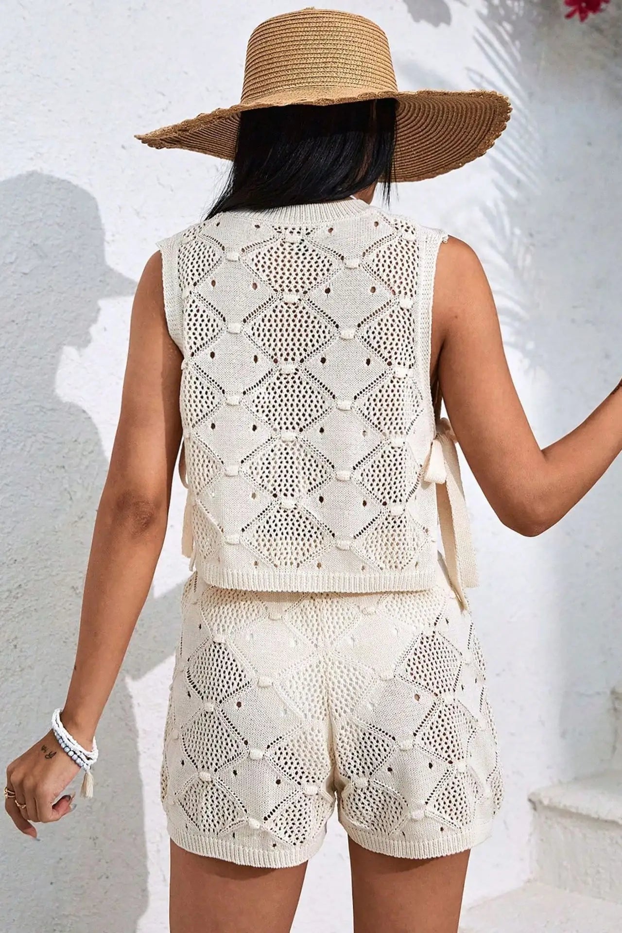 Person wearing a white crochet outfit with a wide-brimmed hat against a white wall.