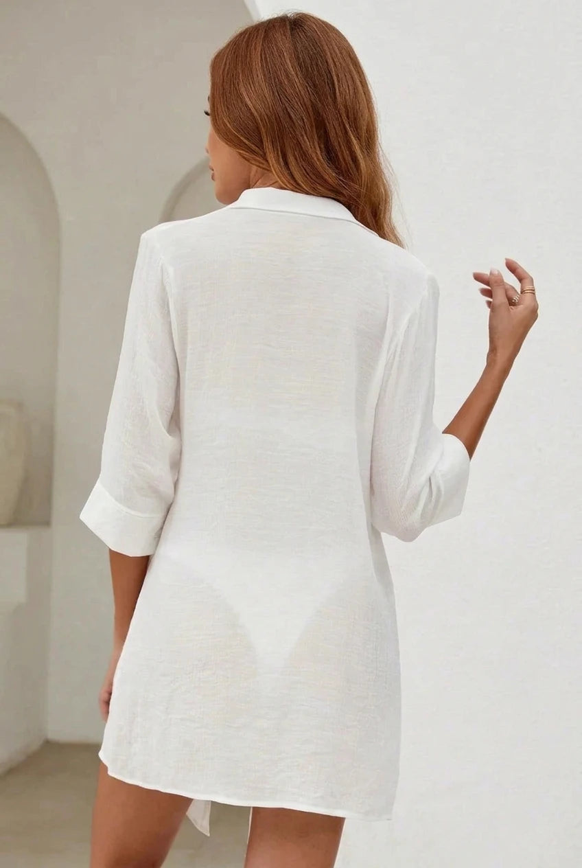 A woman shown from behind wearing a white linen shirt dress with three-quarter sleeves in a bright, minimalist white interior.