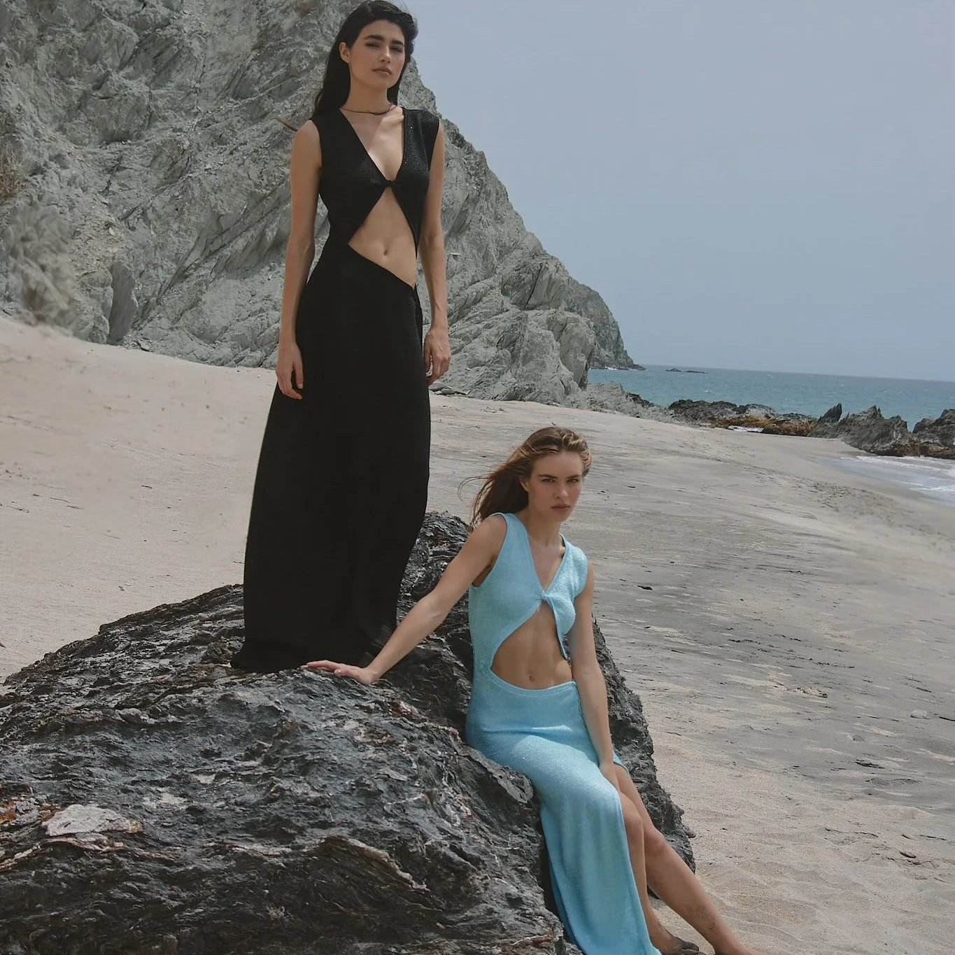 Two women in black and blue dresses on a rocky beach with ocean and sky in the background