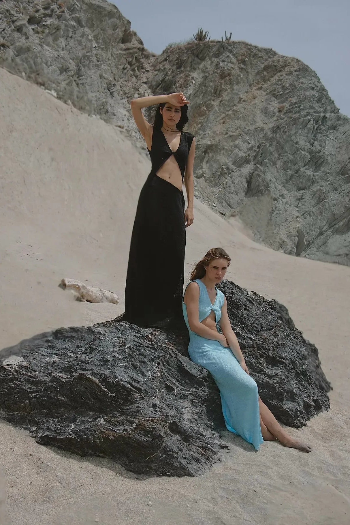 Two women in black and blue dresses standing on a rocky beach with mountains in the background.