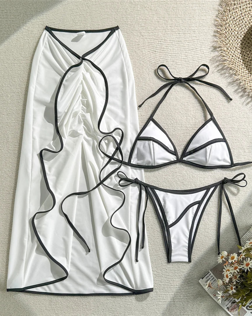 White bikini set with black straps on a textured surface with a hat and flowers.
