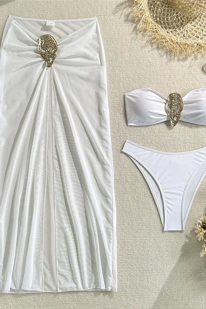White bikini set with gold embellishments and a matching white skirt on a beige surface.