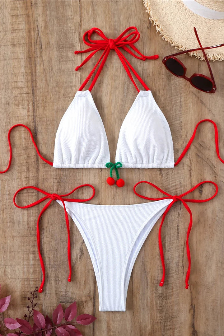 White bikini set with red ties and cherry details on a wooden surface, accompanied by a hat and sunglasses.