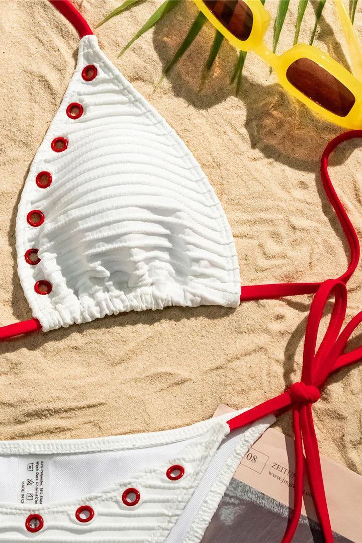 White bikini top with red straps and buttons on sand, with sunglasses in the background.