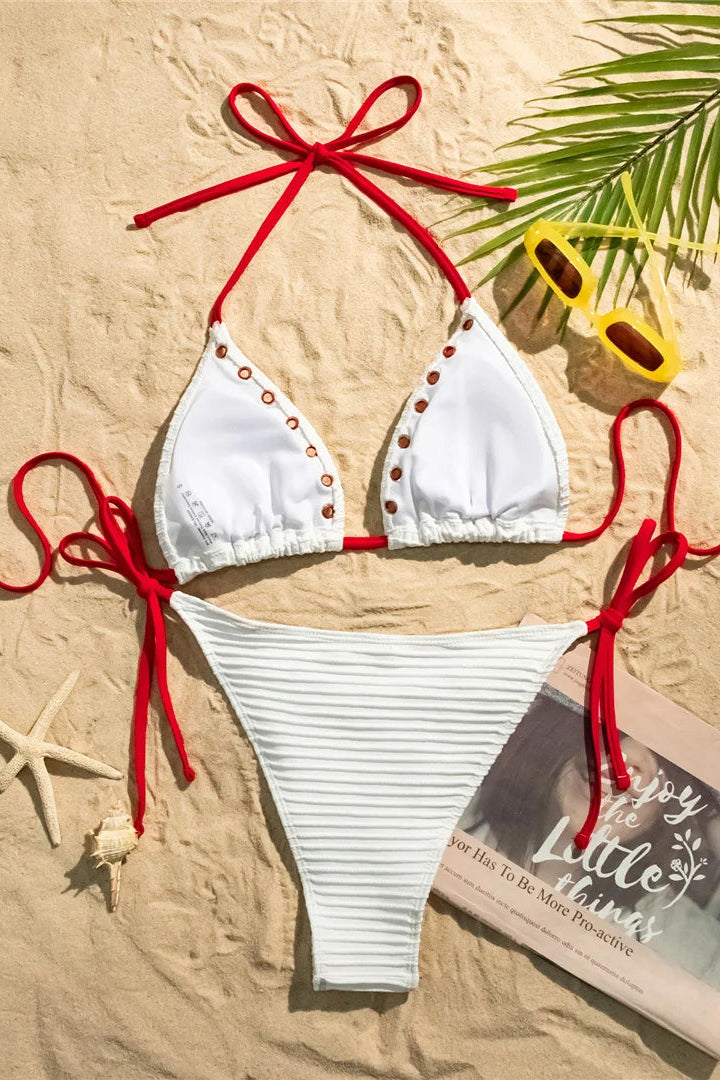 White bikini with red straps on a sandy background with sunglasses and a magazine.