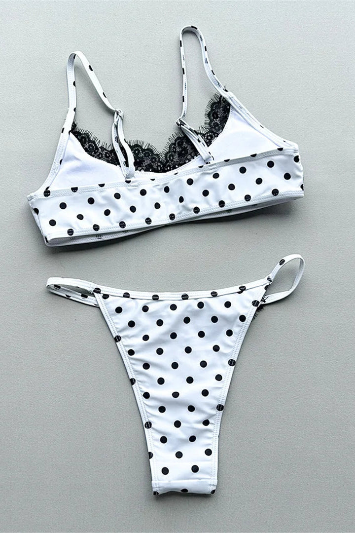 White two-piece bikini set with black polka dots, featuring a bandeau-style bikini top with black lace and matching high-impact thong bottoms, laid flat on a neutral gray background.