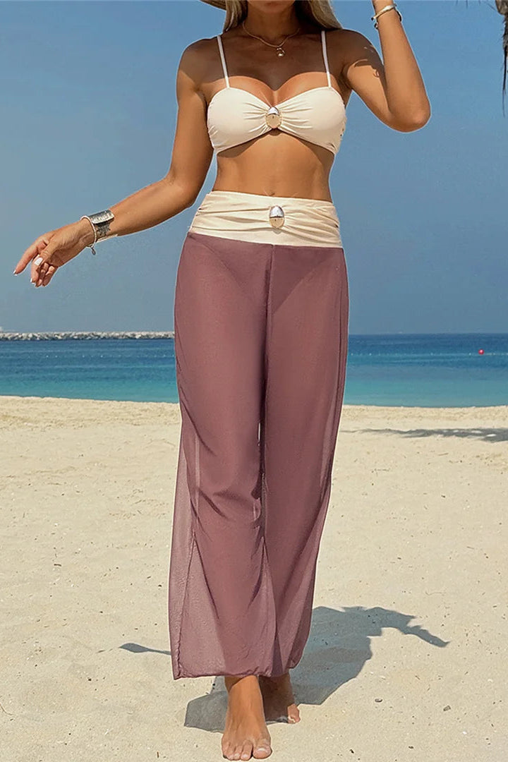Woman in a beige bikini top and mauve pants standing on a beach with palm trees and blue sky.