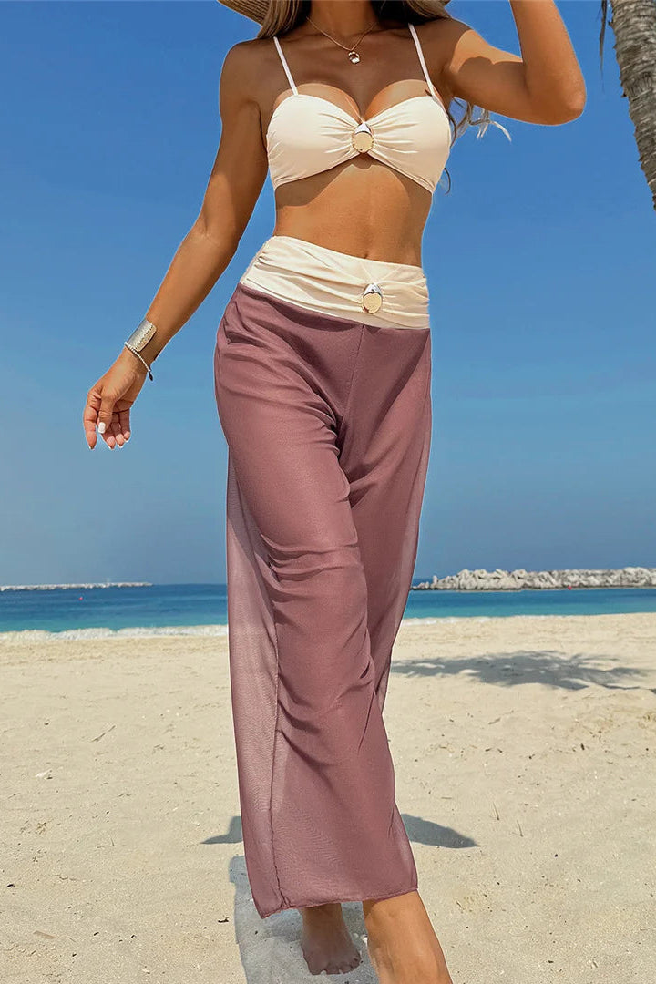 Woman in a beige bikini top and purple pants standing on a beach with palm trees and clear blue sky.