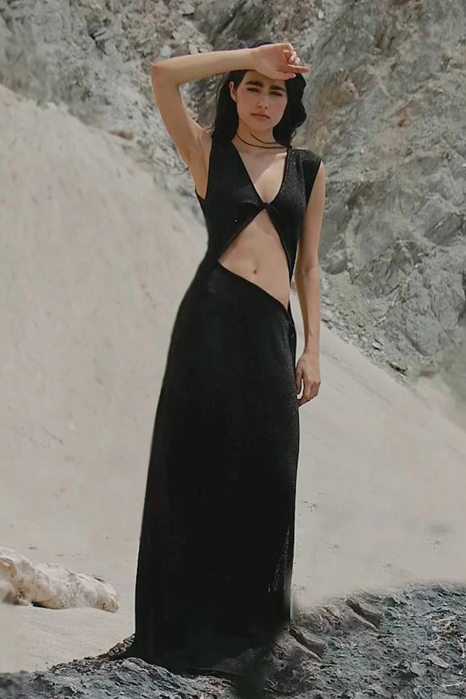 Woman in a black cut out beach dress standing on a rocky beach.