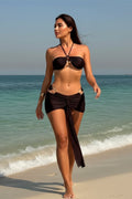 Woman in a brown bikini and sarong standing on a beach with ocean in the background