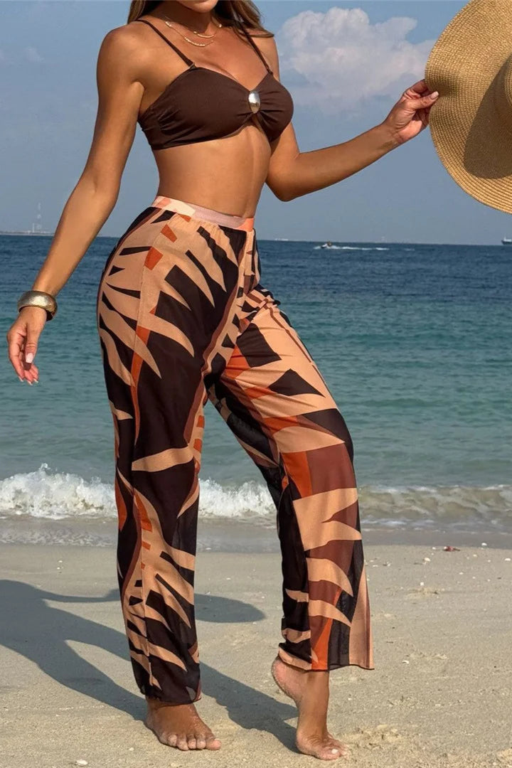 Woman in a brown bikini top and patterned cover up pants standing on a beach with ocean in the background