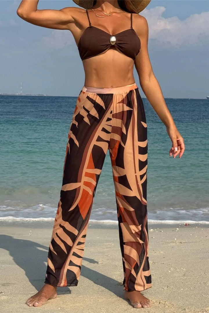 Woman in a brown bikini top and patterned pants standing on a beach with ocean in the background