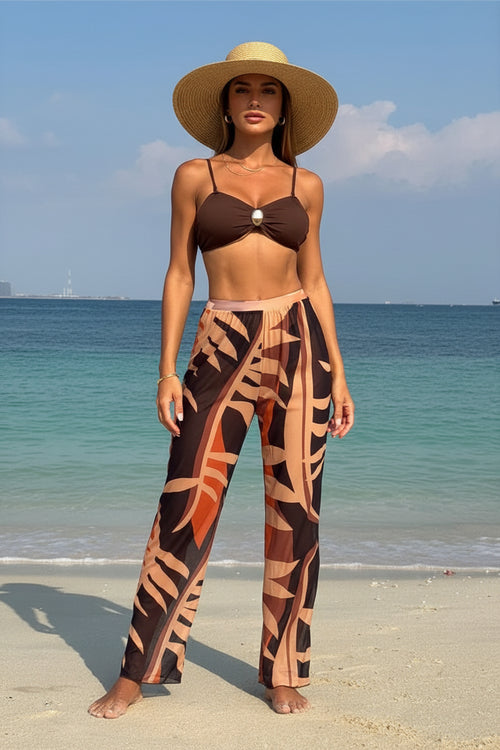 Woman in a brown bikini top and patterned pants standing on a beach with ocean in the background