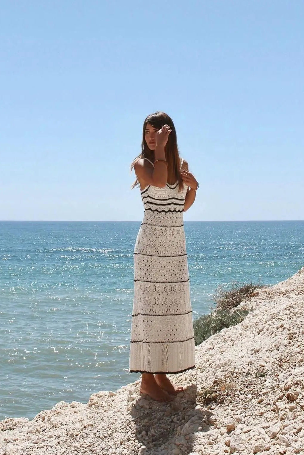 Woman in a dress standing on a rocky cliff overlooking the ocean with clear blue sky.