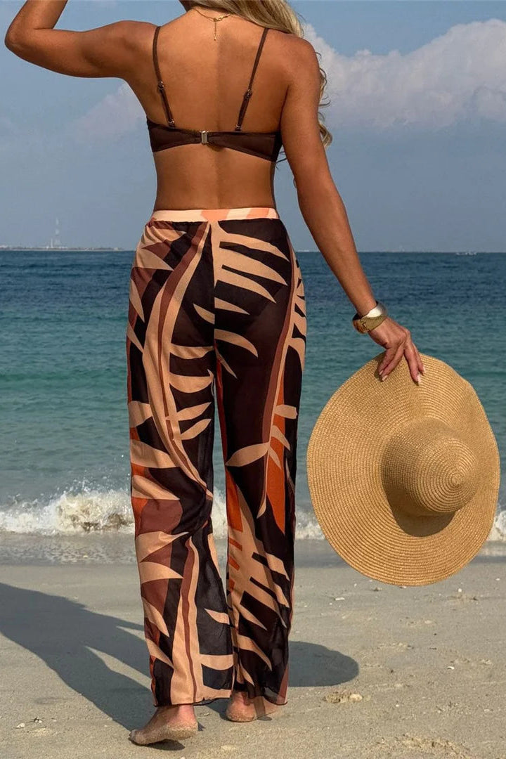 Woman in a leaf-patterned bikini set cover up pants holding a straw hat on a beach.