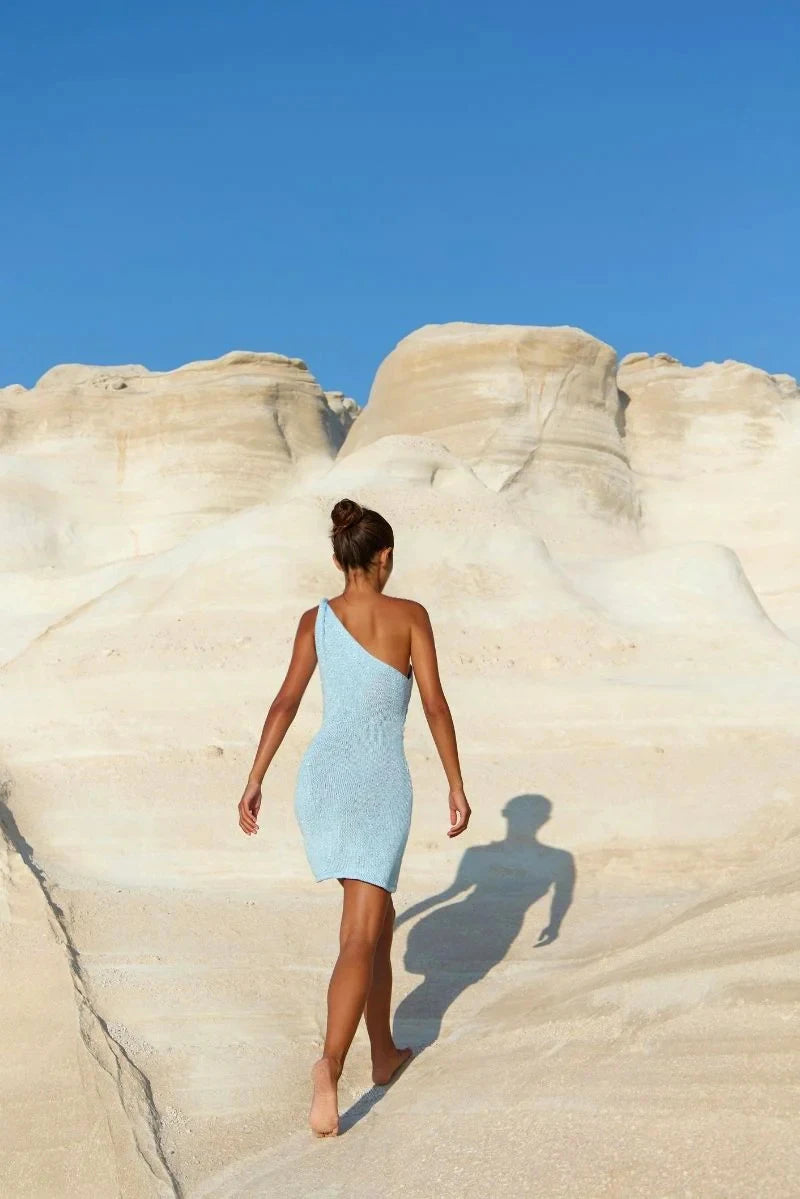 Woman in a light blue dress standing on a rocky beach with a clear blue sky.