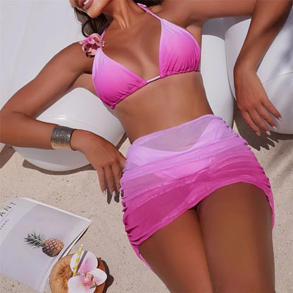 Woman in a pink bikini sitting on a white chair with a magazine and flower on the sand.