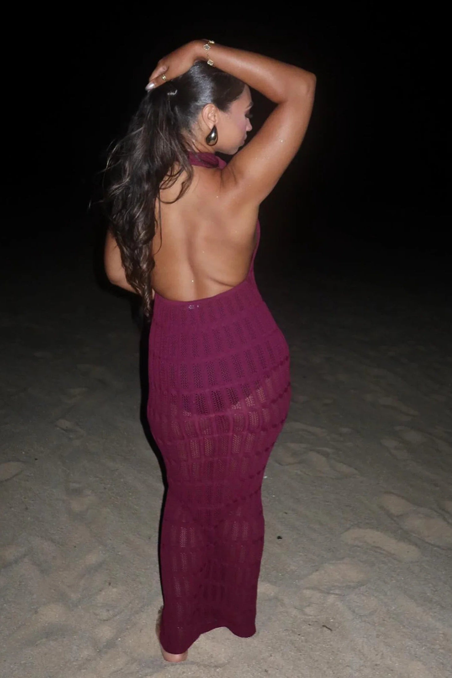 Woman in a purple back less cover up dress standing on a sandy surface at night.