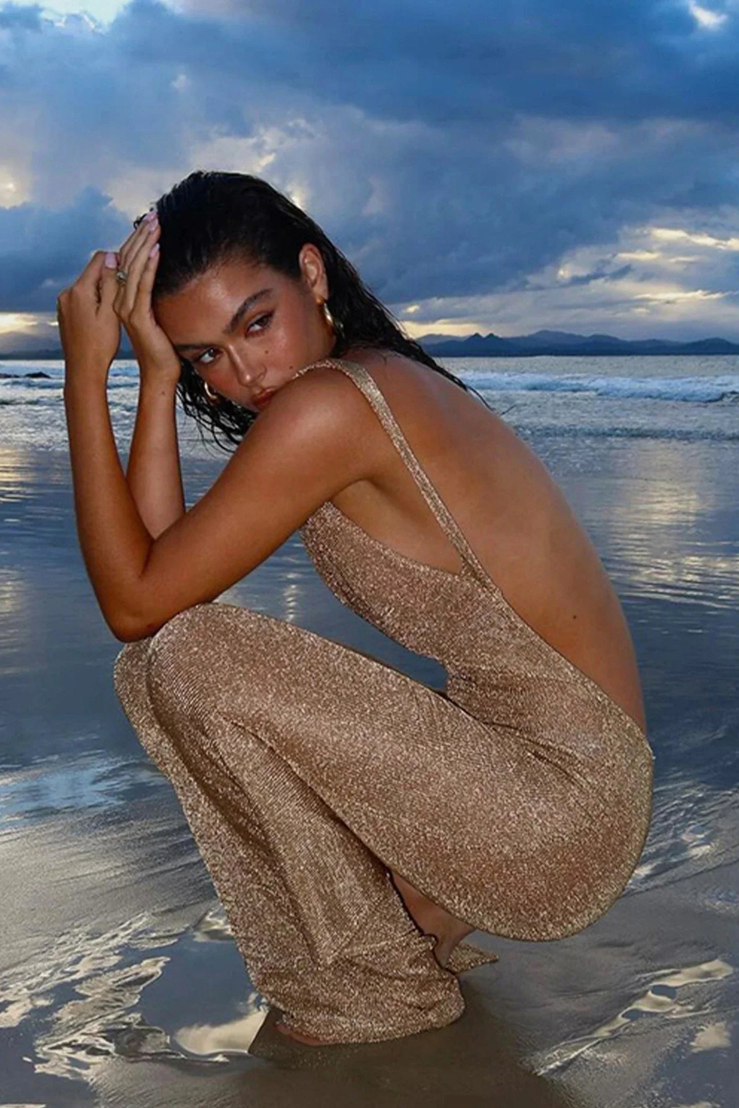 Woman in a sparkly cover up squatting on a beach with a dramatic sky.