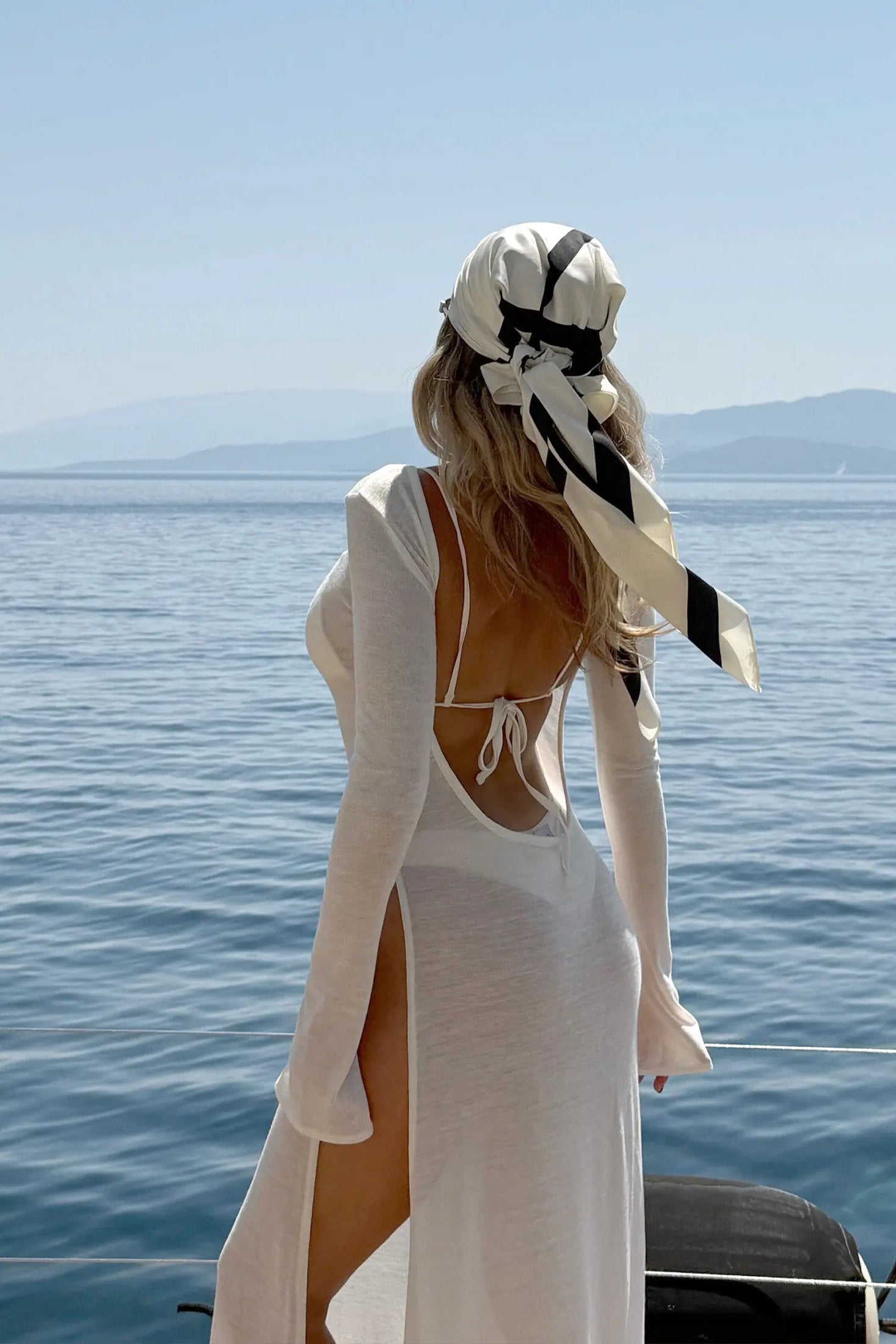Woman in a white cover up dress with a high slit standing on a boat with a scenic background