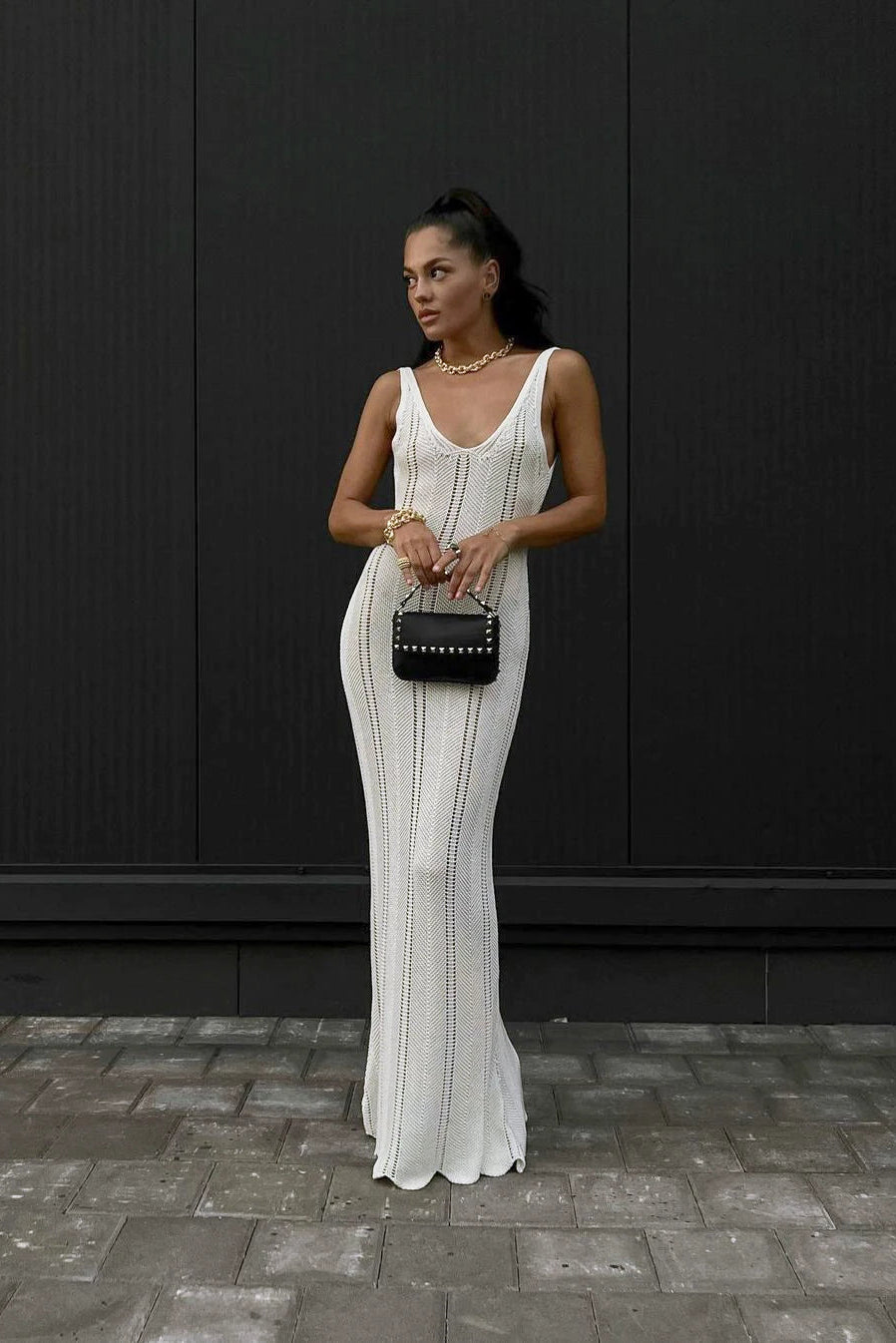 Woman in a white dress holding a black clutch against a dark wall.