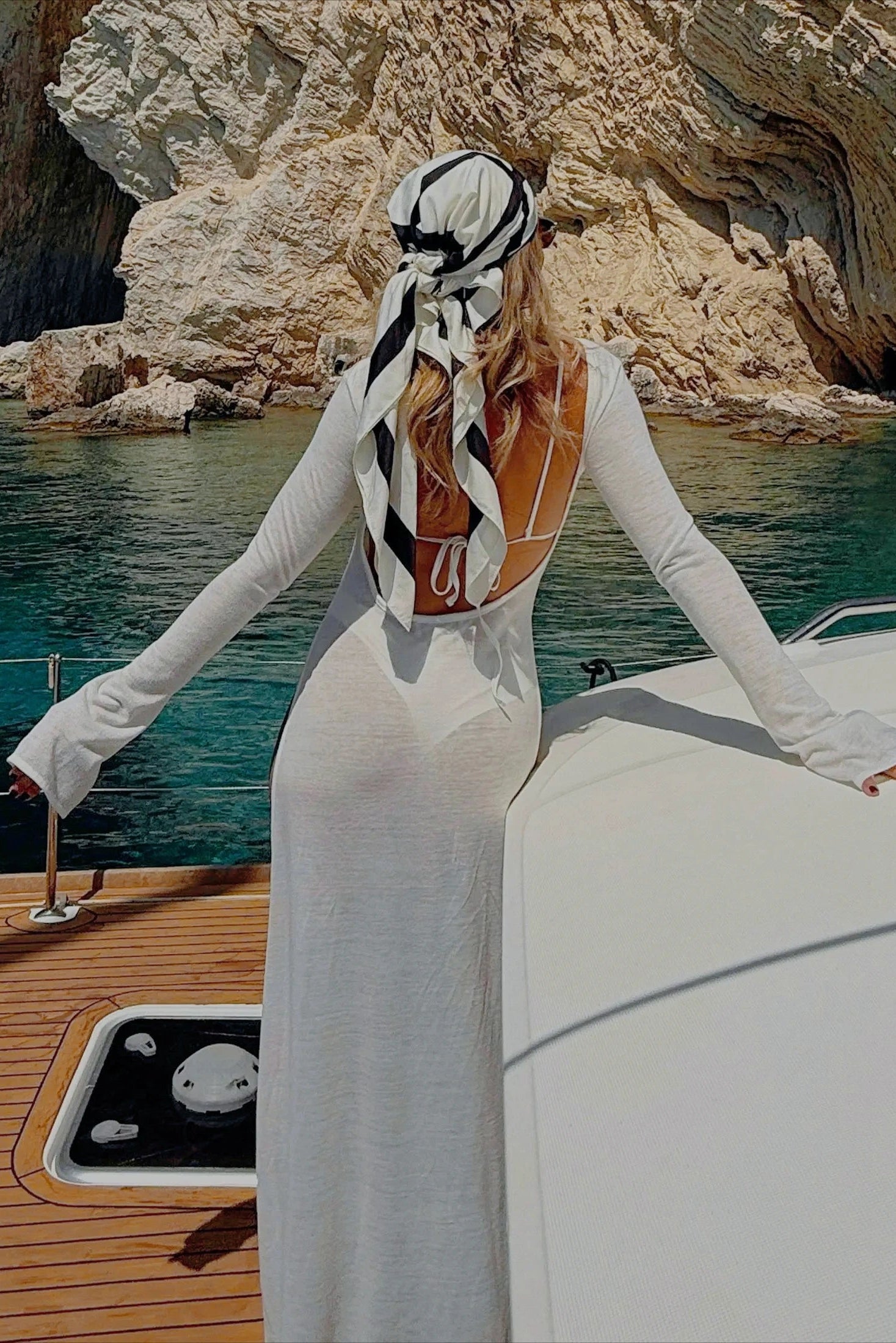 Woman in a white dress with a black and white headscarf on a boat with rocky cliffs in the background