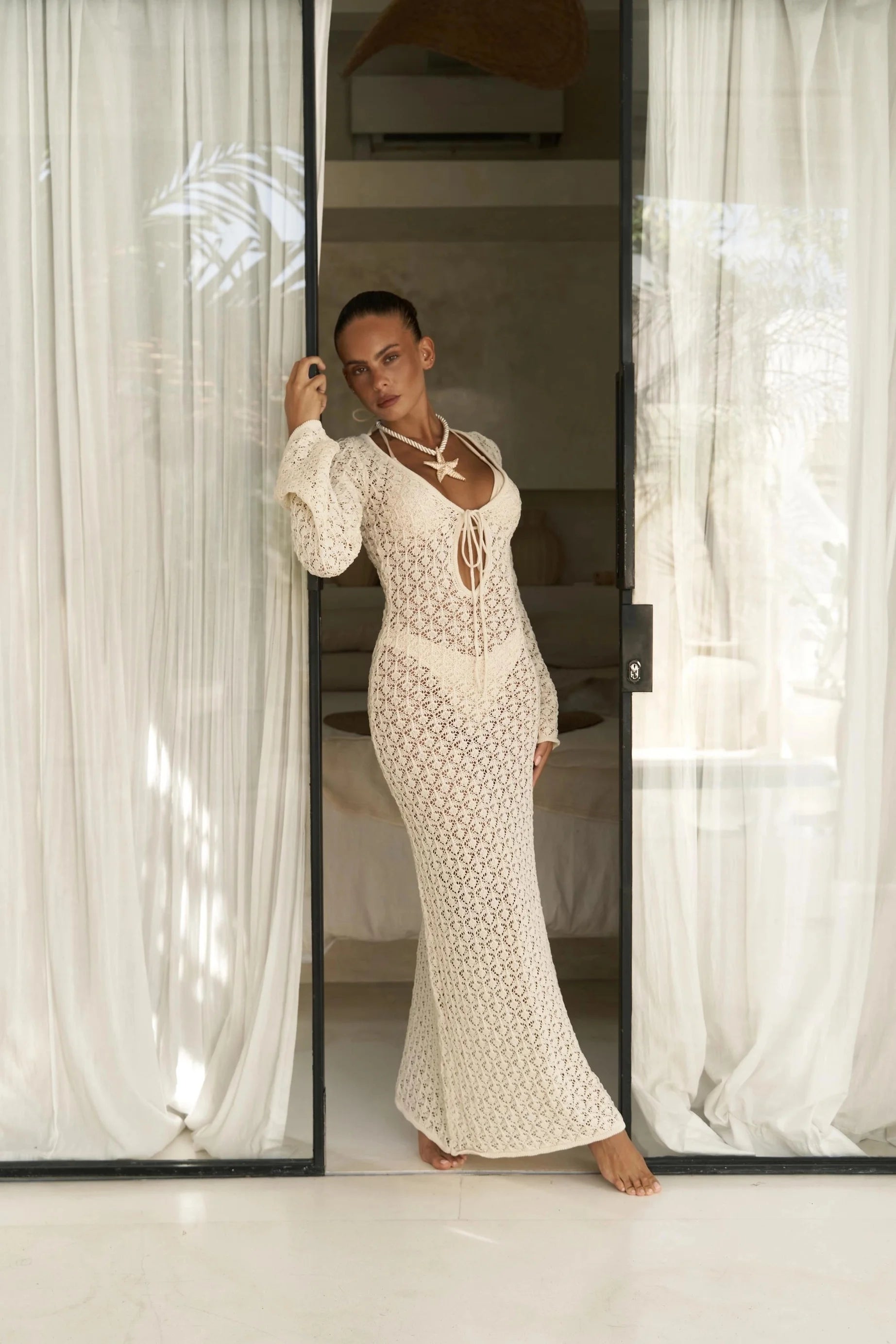 Woman in a white lace beach cover up dress standing in a room with sheer curtains.