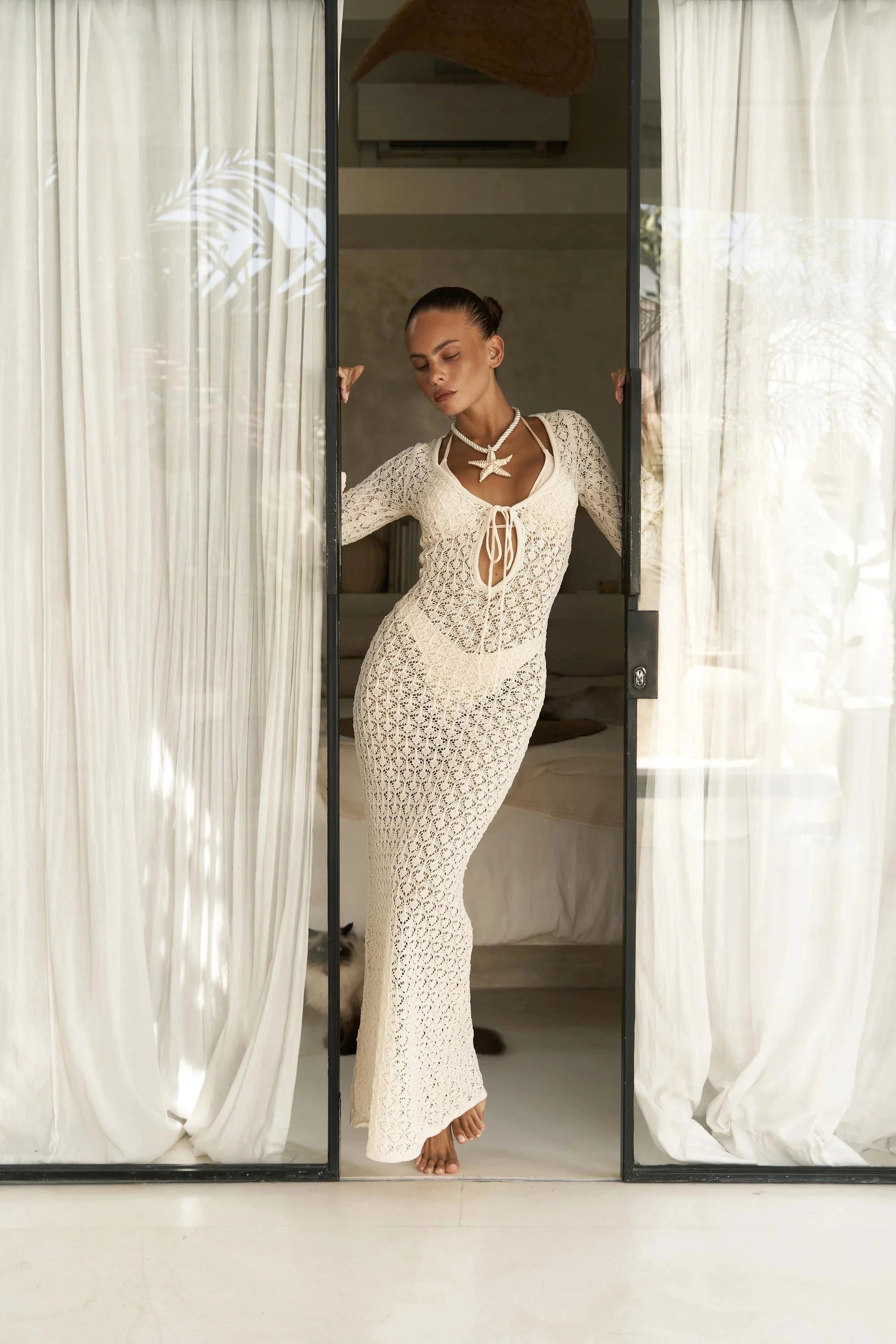 Woman in a white lace cover up dress standing in front of a mirror with sheer curtains.