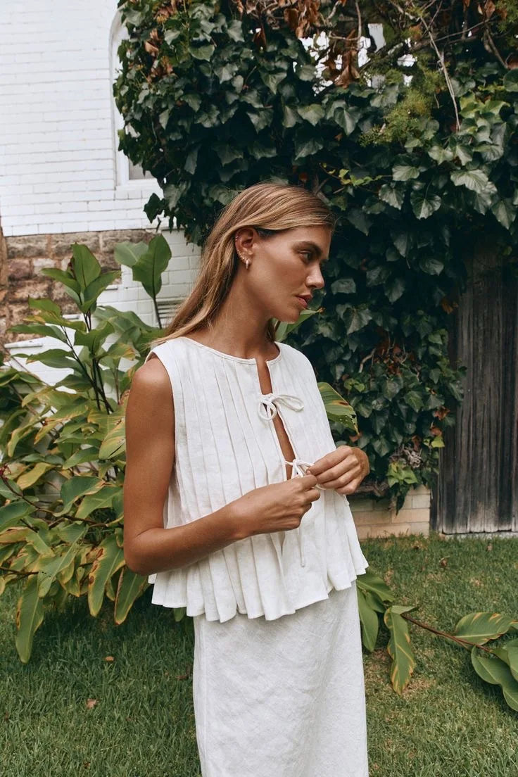 Woman in a white sleeveless top and pants standing outdoors with greenery around