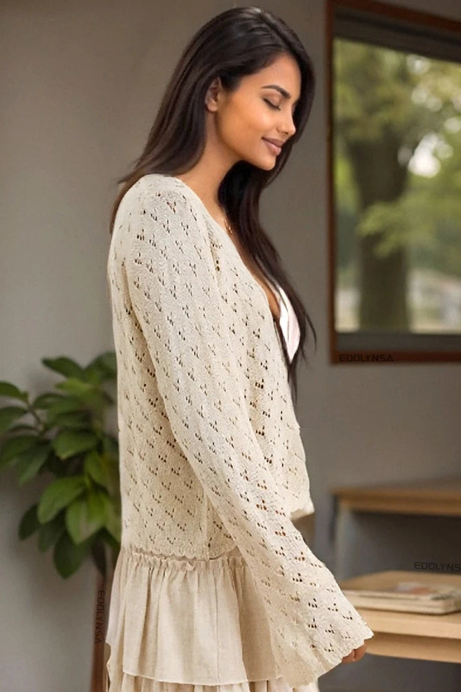 Woman wearing a beige lace cardigan cover up standing indoors with a plant and table in the background
