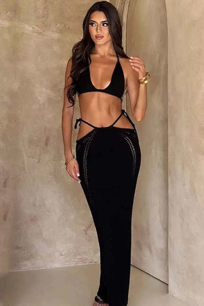 Woman wearing a black beach wear outfit with cut-out design against a neutral wall.