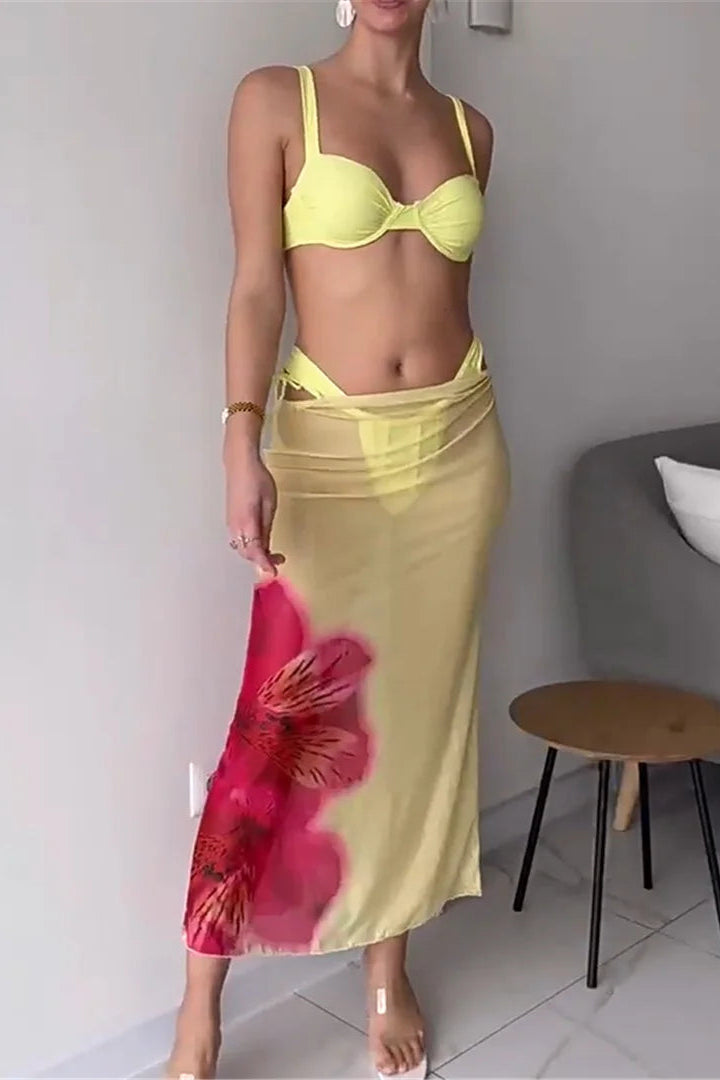 Woman wearing a yellow bikini and floral skirt in a room with a gray wall and white floor.